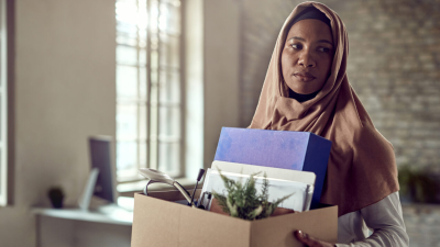 A photo of a woman packing up her things in an office, she is holding a box of her belongings.