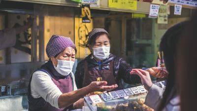 A photograph of two elderly Japanese ladies selling food at a market. They are both wearing face masks.